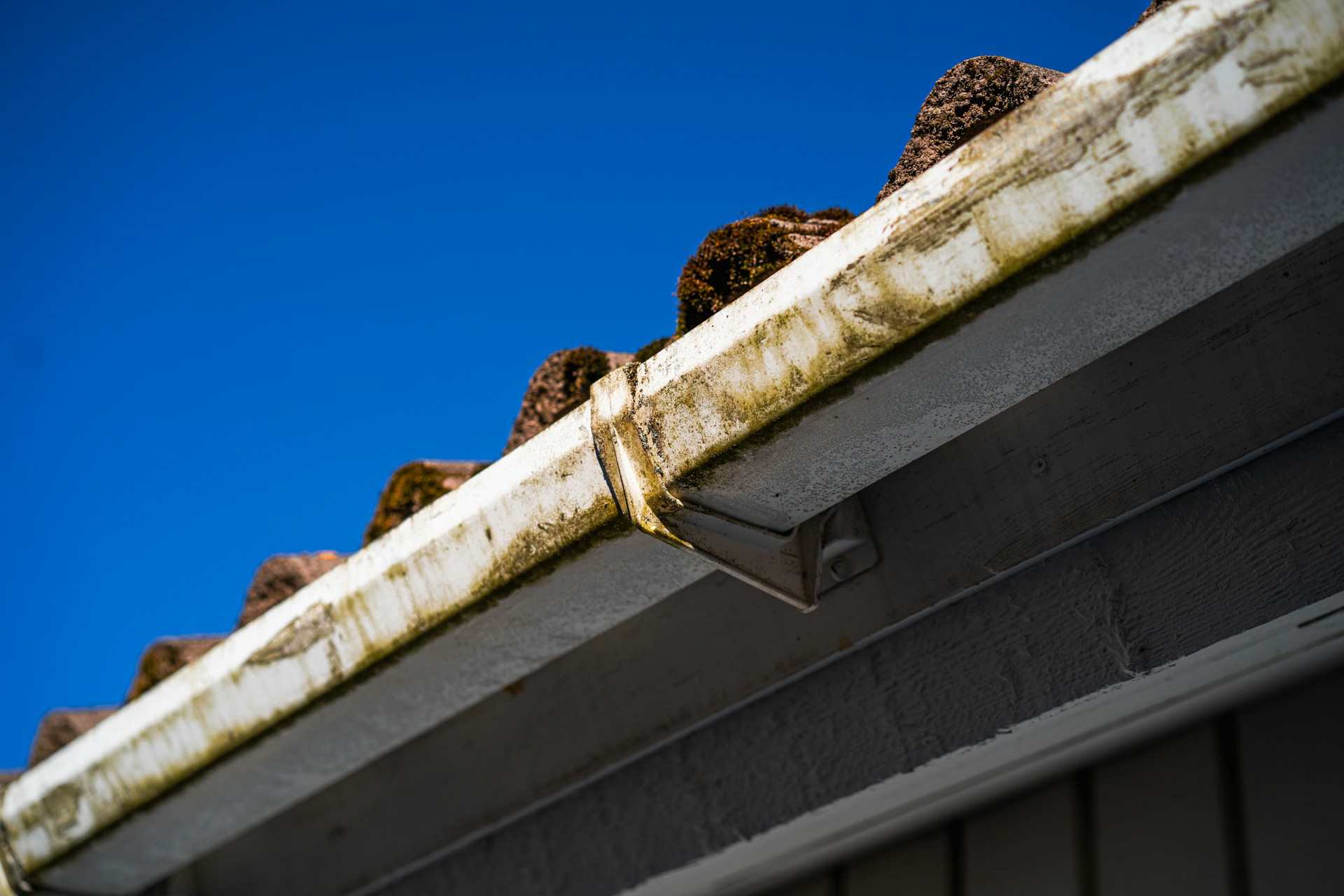 A dirty house gutter attached to the edge of a shingled roofline against a blue sky, illustrating the need for exterior home maintenance.